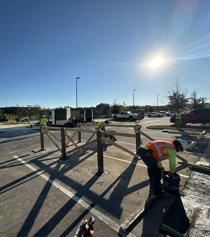 Timber posts braced upright in cored holes in an existing striped asphalt parking lot, crew working alongside